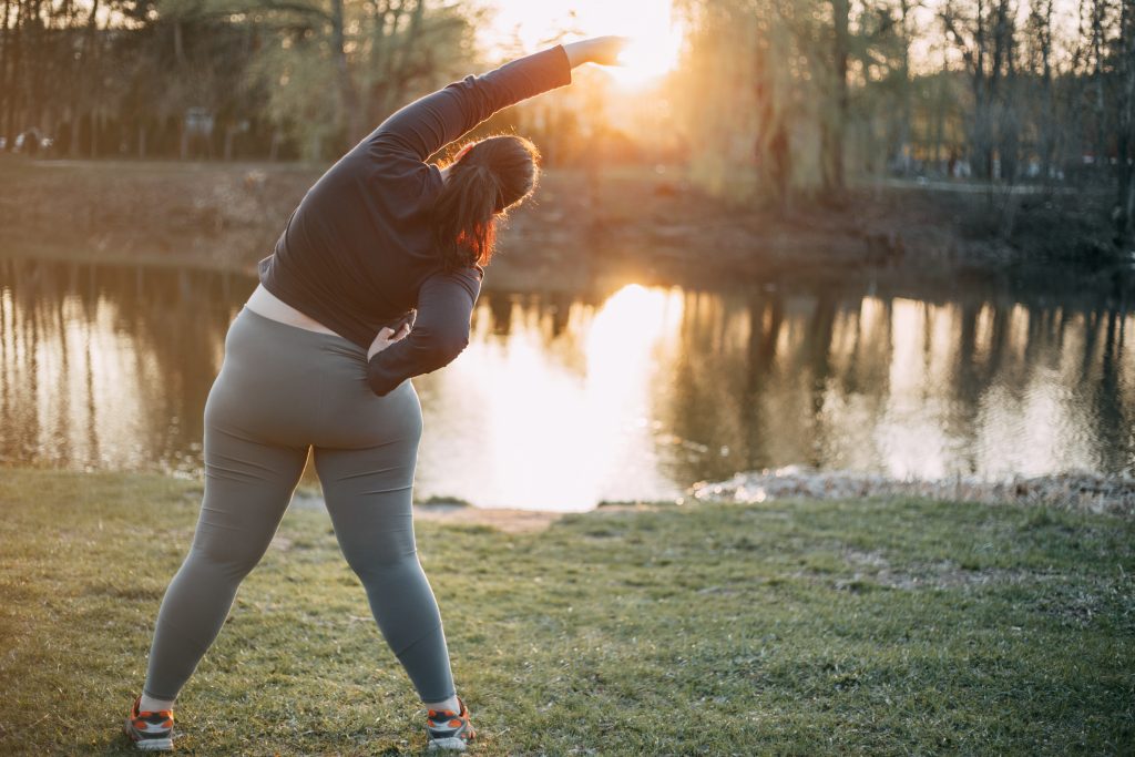 Overweight woman exercising in front of water with sun beaming in the background.